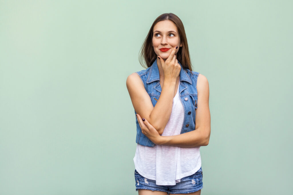 Happy cheerful teenage girl with freckles, casual style white t shirt and jeans jacket, looking up, thinking and touching her face and a, enjoying good day and free time indoors. Studio shot, light green background