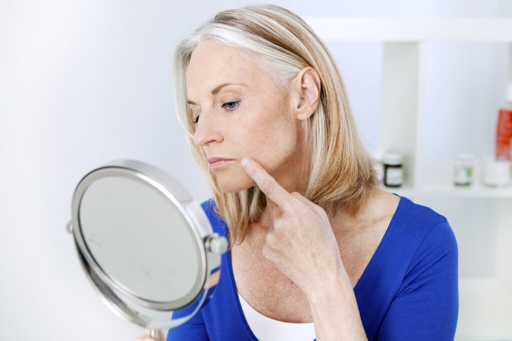 Woman in blue examining her marionette lines in a handheld mirror.